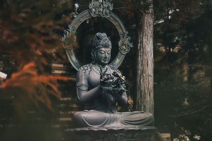 A dark bronze statue of a seated Buddha holding a flower, surrounded by green forest trees. | PrintPosters.in