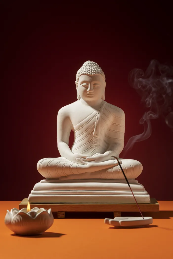 A white Lord Buddha statue in meditation pose with burning incense and a lit lotus candle against a red background. | PrintPosters.in