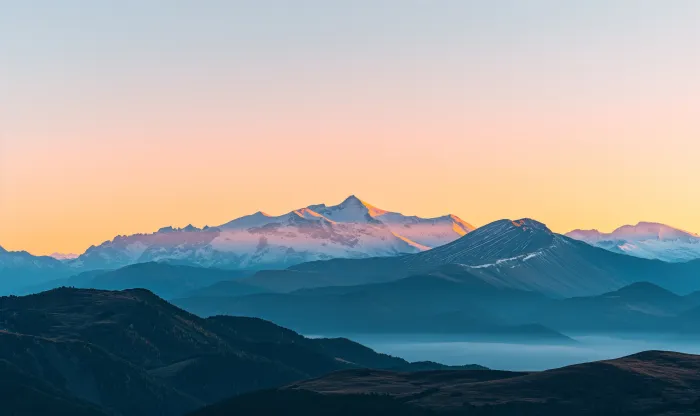 Panoramic view of majestic snow-capped mountain peaks illuminated by golden sunset light with misty blue ridges below. | PrintPosters.in