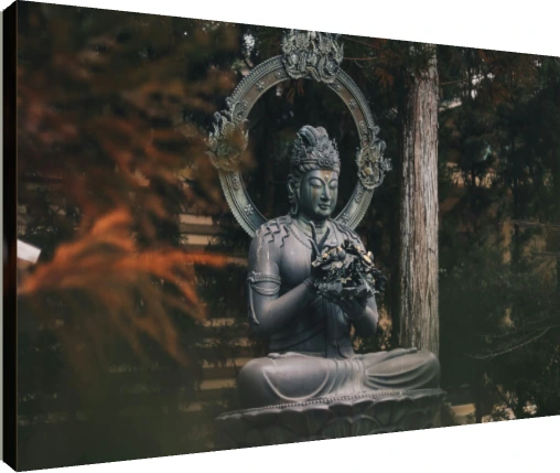 A dark bronze statue of a seated Buddha holding a flower, surrounded by green forest trees. | PrintPosters.in